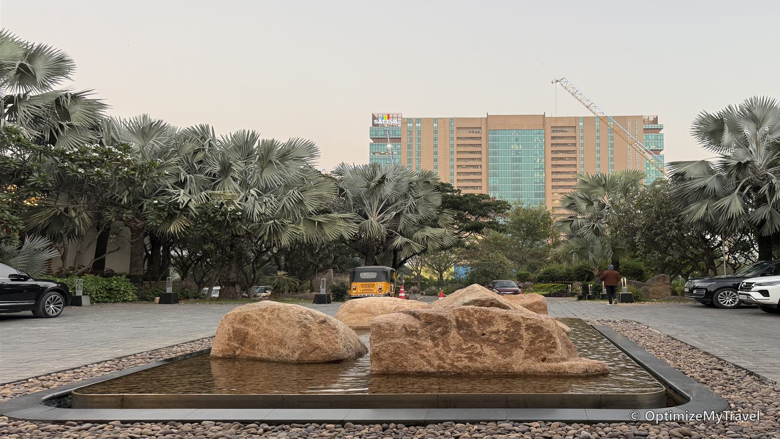 Hotel arrival drive with parked cars, landscaped rocks, leafy trees, and an office tower beyond